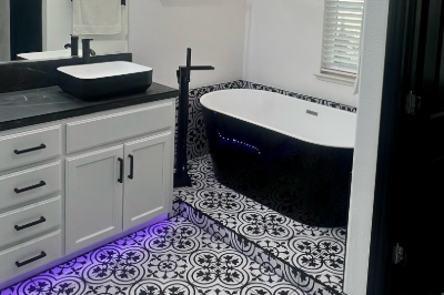 Modern bathroom renovation with black and white patterned tile flooring, white cabinetry, a sleek black countertop, dual black vessel sinks, and a freestanding black bathtub with purple LED accent lighting.